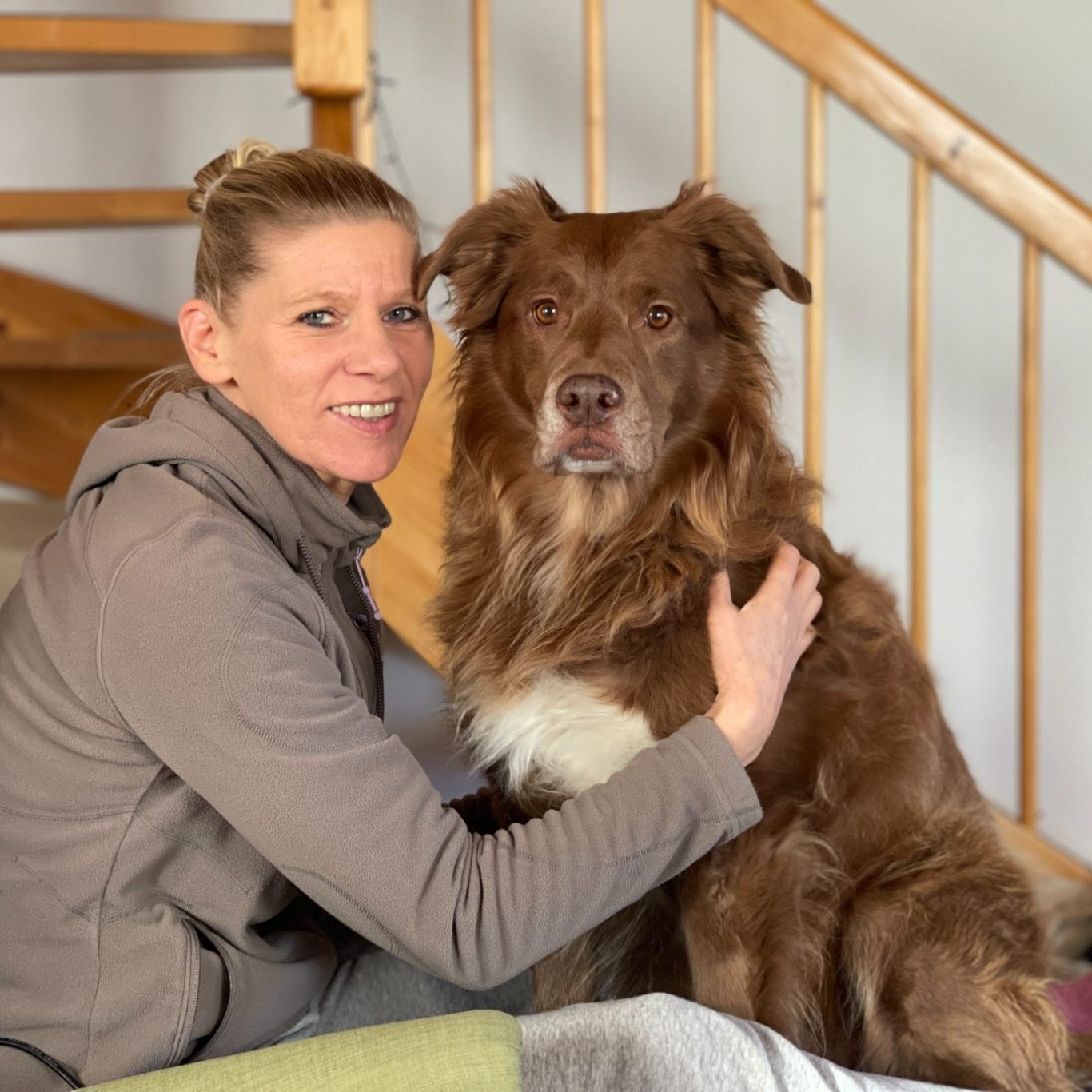 Eine Frau mit einem braunen Hund sitzt auf einem Sofa in einem hellen Raum.